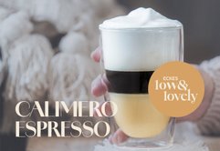 Ein elegantes Glas mit Schichten aus Espresso, cremigem Schaum und goldener Flüssigkeit wird von einer Hand gehalten.