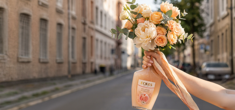 Eine Hand hält eine ECKES Weißer Pfirsich Flasche mit einem Blumenstrauß aus  cremefarbenen und apricotfarbenen Blumen. Die Flasche dient als Vase vor einer  Straße mit Gebäuden im Hintergrund.