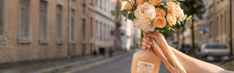 Eine Hand hält eine ECKES Weißer Pfirsich Flasche mit einem Blumenstrauß aus  cremefarbenen und apricotfarbenen Blumen. Die Flasche dient als Vase vor einer  Straße mit Gebäuden im Hintergrund.