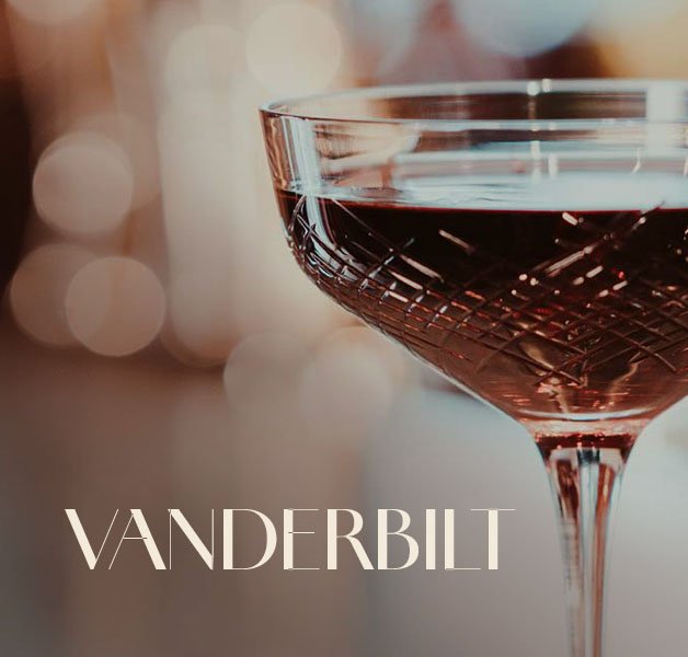 Glas mit ECKES Vanderbilt Cocktail, vor einem unscharfen, warm beleuchteten Hintergrund mit der Aufschrift "Vanderbilt"