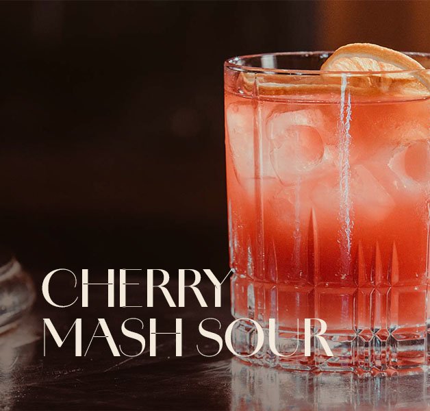 Glas mit ECKES Cherry Mash Sour Cocktail auf Eis, garniert mit einer Orangenscheibe, vor einem dunklen Hintergrund