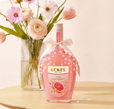 Mit Perlen und einer Schleife dekorierte ECKES Pink Grapefruit Flasche auf einem  Holztisch vor einer Glasvase mit rosa und weißen Blumen. Einige Perlen liegen  auf dem Tisch.