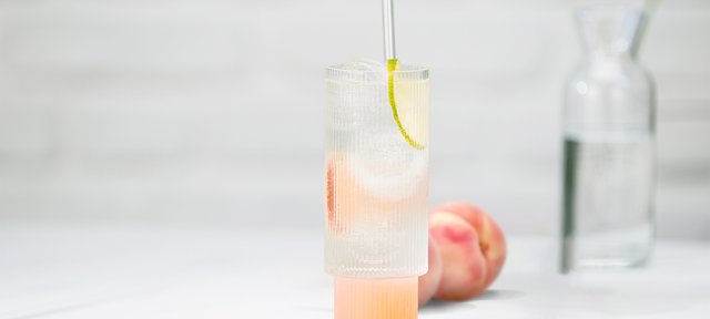 Glas mit ECKES Peach Tonic Cocktail auf Eis, garniert mit einer Zitronenscheibe, vor einem weißen Hintergrund mit einem frischen Pfirsich.