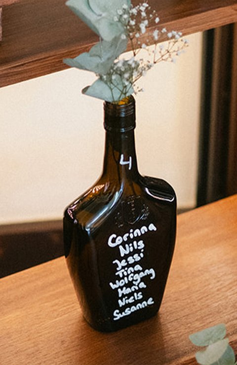 Eine beschriftete dunkle ECKES Flasche mit weißen Blumen und  Eukalyptus dient als Sitzplan für eine Hochzeit auf einem mehrstufigen  Holzregal.