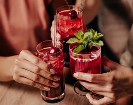 Mehrere Hände stoßen mit drei roten Cocktails in Tumblergläsern an. Die Drinks  sind mit Kirschen, Minze und Glasstrohhalmen garniert.