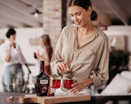 Eine Frau in einem beigen Hemd dekoriert zwei Cocktails mit Minze und Kirschen auf einem Holzbrett.
