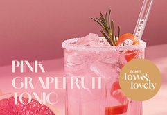 Ein erfrischendes Glas pinker Grapefruit-Tonic mit Eiswürfeln und einer dekorativen Orangenschale auf einem rosa Hintergrund.