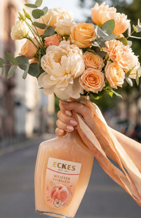 Eine Hand hält eine ECKES Weißer Pfirsich Flasche mit einem Blumenstrauß aus  cremefarbenen und apricotfarbenen Blumen. Die Flasche dient als Vase vor einer  Straße mit Gebäuden im Hintergrund.