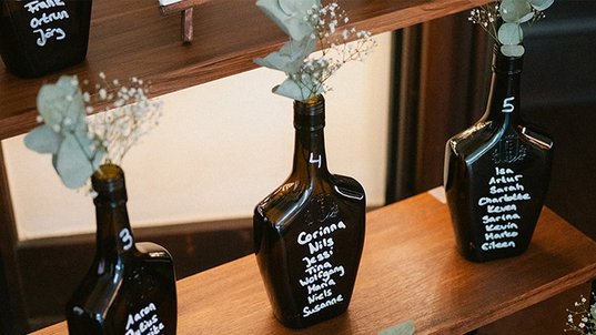 Mehrere beschriftete dunkle ECKES Flaschen mit weißen Blumen und  Eukalyptus dienen als Sitzplan für eine Hochzeit auf einem mehrstufigen  Holzregal.