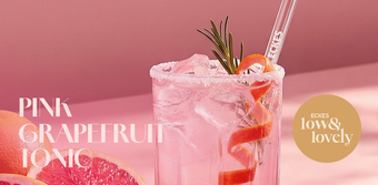 Ein erfrischendes Glas pinker Grapefruit-Tonic mit Eiswürfeln und einer dekorativen Orangenschale auf einem rosa Hintergrund.