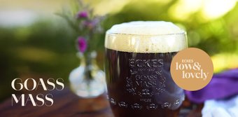 Im Vordergrund steht ein Glas mit schaumigem, dunklem Bier, während im Hintergrund sanfte, grüne Farben und Blumen sichtbar sind.