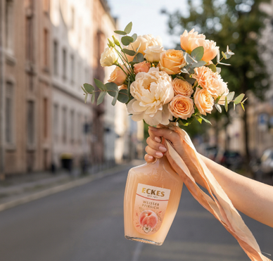 Eine Hand hält eine ECKES Weißer Pfirsich Flasche mit einem Blumenstrauß aus  cremefarbenen und apricotfarbenen Blumen. Die Flasche dient als Vase vor einer  Straße mit Gebäuden im Hintergrund.