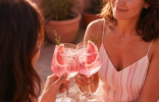 Zwei Frauen stoßen mit rosafarbenen Cocktails, Grapefruitscheiben und  Kräutergarnitur in Weingläsern an.