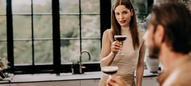 In einer stilvollen Küche genießt eine Frau einen Cocktail und strahlt Selbstbewusstsein aus, während sie mit einem Mann spricht.