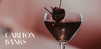 Ein elegantes Cocktailglas mit dunkler Flüssigkeit und einer Kirsche, das auf einem stilvollen Tisch steht.