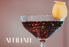 Ein elegantes Cocktailglas mit dunklem Getränk und einer Zitronenschale als Garnitur, vor einem sanften Hintergrund.