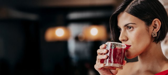 Die Frau mit dem kurzen Haar und dem roten Lippenstift hebt ein Glas mit einem fruchtigen Getränk an, während sie in die Ferne blickt.