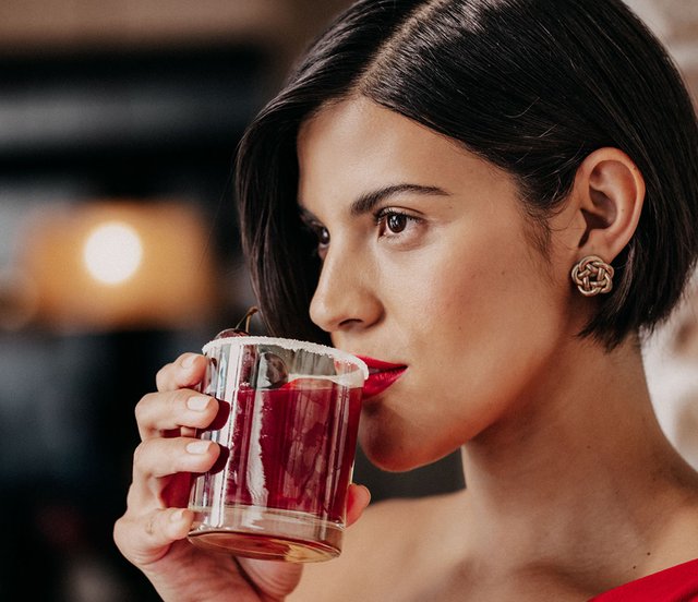 Die Frau mit dem kurzen Haar und dem roten Lippenstift hebt ein Glas mit einem fruchtigen Getränk an, während sie in die Ferne blickt.