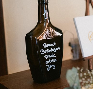 Eine beschriftete dunkle ECKES Flasche mit weißen Blumen und Eukalyptus dient als Sitzplan für eine Hochzeit auf einem mehrstufigen Holzregal.