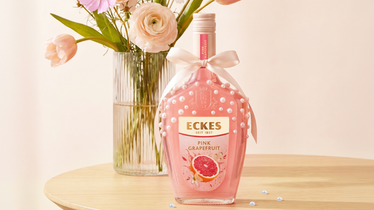 Mit Perlen und einer Schleife dekorierte ECKES Pink Grapefruit Flasche auf einem  Holztisch vor einer Glasvase mit rosa und weißen Blumen. Einige Perlen liegen  auf dem Tisch.