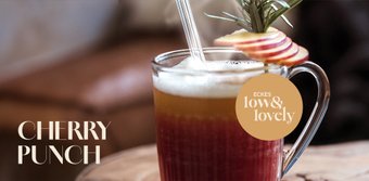 Ein Glas mit Kirschpunsch, dekoriert mit Apfelscheiben und einem Rosmarinzweig, auf einem Holztisch platziert.