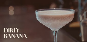 Ein elegantes Cocktailglas mit einem cremigen, hellbraunen Getränk steht auf einer Bar, umgeben von verschwommenen Lichtern.
