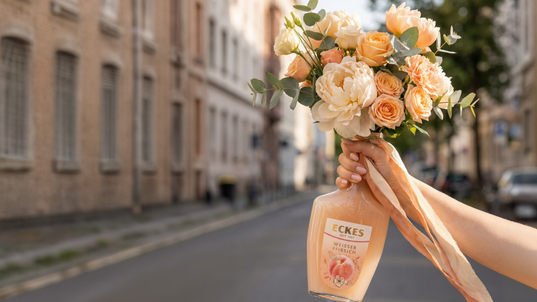Eine Hand hält eine ECKES Weißer Pfirsich Flasche mit einem Blumenstrauß aus  cremefarbenen und apricotfarbenen Blumen. Die Flasche dient als Vase vor einer  Straße mit Gebäuden im Hintergrund.
