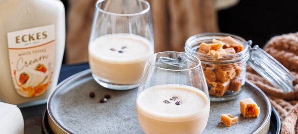 Ein rustikales Tablett zeigt zwei Gläser mit Kaffee-Creme, umgeben von Kaffeebohnen und einer Glasdose voller Toffee-Stücke.