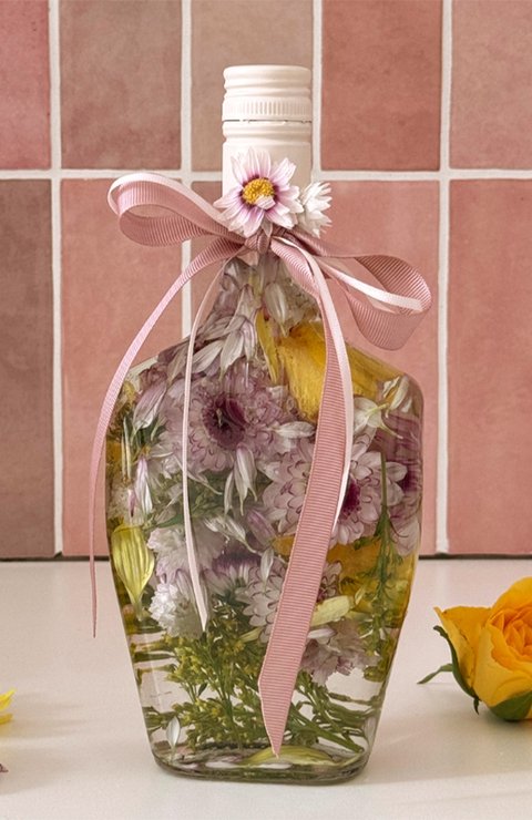 Mit Blumen, transparentem Öl und einer rosafarbenen Schleife dekorierte ECKES  Flasche vor rosafarbenen Fliesen. Daneben liegen gelbe und lilafarbene Blumen.