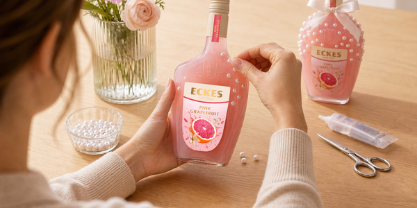 Eine Person verziert eine ECKES Pink Grapefruit Flasche mit selbstklebenden  Perlen. Auf dem Tisch liegen eine Schale mit Perlen, eine Schere und Kleber. Im  Hintergrund steht eine fertig dekorierte Flasche mit Schleife.