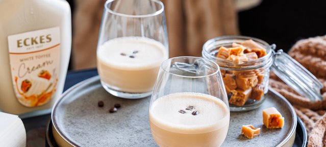 Ein rustikales Tablett zeigt zwei Gläser mit Kaffee-Creme, umgeben von Kaffeebohnen und einer Glasdose voller Toffee-Stücke.