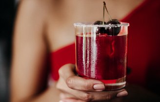 Ein erfrischendes Getränk in einem Glas, verziert mit Kirschen, wird von einer Hand gehalten, während der Hintergrund verschwommen ist.