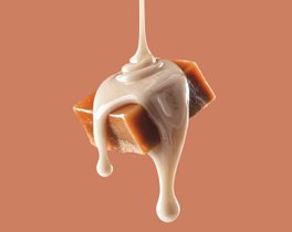 Ein Stück Karamell, überzogen mit einer cremigen, hellen Sauce, die langsam herab tropft, vor einem warmen, einfarbigen Hintergrund.