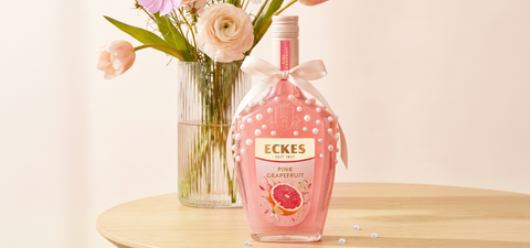 Mit Perlen und einer Schleife dekorierte ECKES Pink Grapefruit Flasche auf einem  Holztisch vor einer Glasvase mit rosa und weißen Blumen. Einige Perlen liegen  auf dem Tisch.