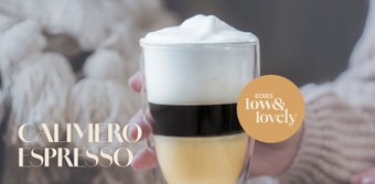 Ein elegantes Glas mit Schichten aus Espresso, cremigem Schaum und goldener Flüssigkeit wird von einer Hand gehalten.