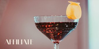 Ein elegantes Cocktailglas mit dunklem Getränk und einer Zitronenschale als Garnitur, vor einem sanften Hintergrund.