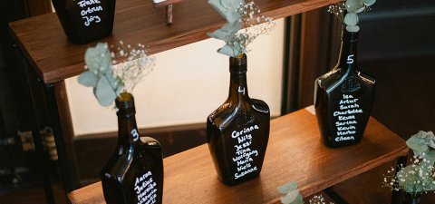 Mehrere beschriftete dunkle ECKES Flaschen mit weißen Blumen und  Eukalyptus dienen als Sitzplan für eine Hochzeit auf einem mehrstufigen  Holzregal.