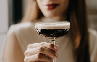 Eine Frau mit langen Haaren hält ein elegantes Glas mit dunklem Getränk und einer schaumigen Oberseite in der Hand.