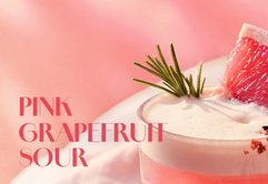 Ein erfrischender pinker Grapefruit-Cocktail, garniert mit einer Grapefruit-Scheibe und Rosmarin, vor einem sanften rosa Hintergrund.
