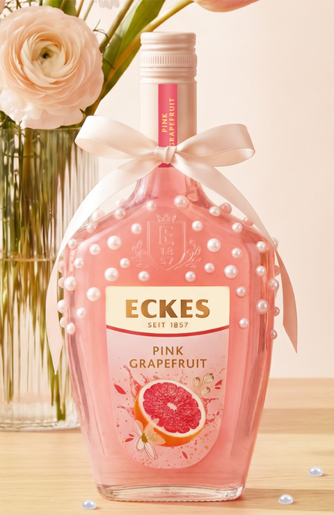 Mit Perlen und einer Schleife dekorierte ECKES Pink Grapefruit Flasche auf einem  Holztisch vor einer Glasvase mit rosa und weißen Blumen. Einige Perlen liegen  auf dem Tisch.