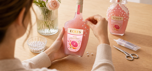Eine Person verziert eine ECKES Pink Grapefruit Flasche mit selbstklebenden  Perlen. Auf dem Tisch liegen eine Schale mit Perlen, eine Schere und Kleber. Im  Hintergrund steht eine fertig dekorierte Flasche mit Schleife.