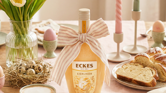 Mit einer Stoffschleife dekorierte ECKES Eierlikör Flasche auf einem gedeckten  Tisch mit Kaffee, Hefezopf, bunten Eiern, Kerzen und Frühlingsblumen.