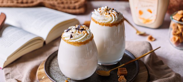 Ein stilvolles Arrangement mit zwei Gläsern, gefüllt mit einem cremigen Getränk, umgeben von Karamellstückchen und einem Buch.