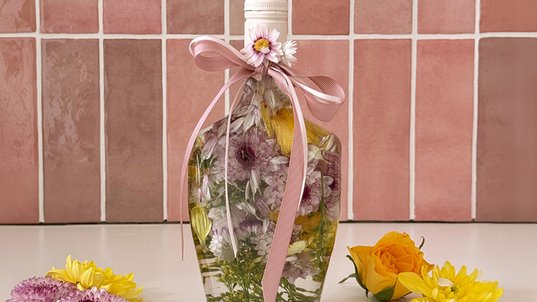 Mit Blumen, transparentem Öl und einer rosafarbenen Schleife dekorierte ECKES  Flasche vor rosafarbenen Fliesen. Daneben liegen gelbe und lilafarbene Blumen.