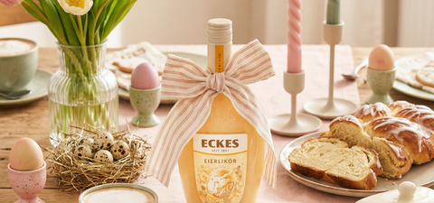 Mit einer Stoffschleife dekorierte ECKES Eierlikör Flasche auf einem gedeckten  Tisch mit Kaffee, Hefezopf, bunten Eiern, Kerzen und Frühlingsblumen.