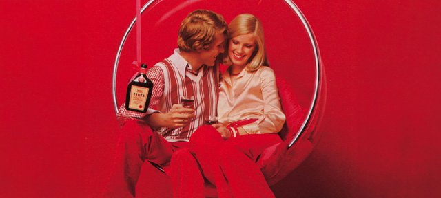 Ein Paar lächelnd vor rotem Hintergrund, der Mann hält eine Flasche ECKES Edelkirsch Likör